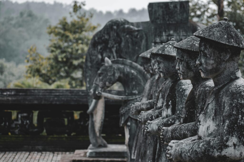 Row of weathered stone statues in a misty, forested area, suggesting historical significance.